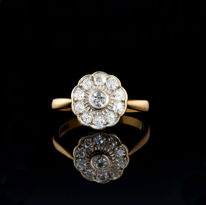 Antique diamond daisy cluster ring with old cut diamonds set in a scalloped platinum topped design on an 18ct gold band, reflecting softly against a dark surface.